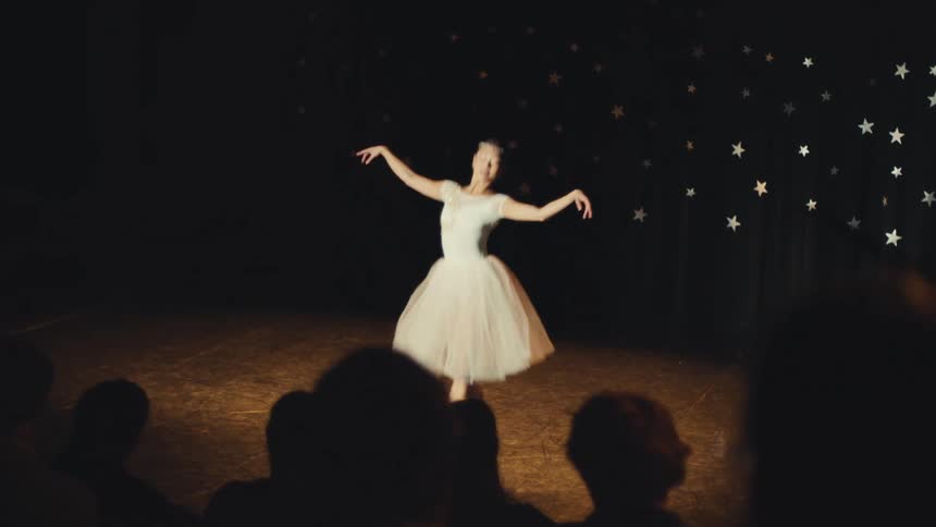 A graceful ballerina in a white tutu performs an elegant classical dance on a dimly lit stage in front of a silhouetted audience