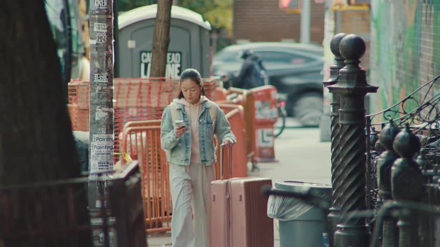 A young Asian woman pulls her suitcase down a city sidewalk, looking up from her phone to find her way in slow motion