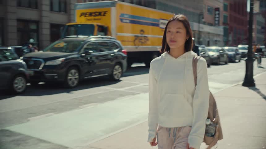 A young Asian woman in a white hoodie walks down a sunny city sidewalk in slow motion