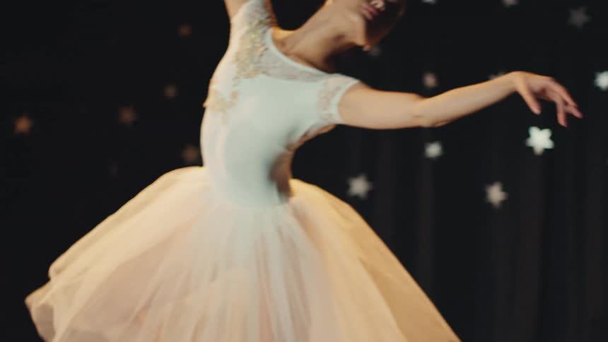 In slow motion, a graceful ballerina in a white tutu and tiara performs an elegant ballet routine on a stage with a starry background