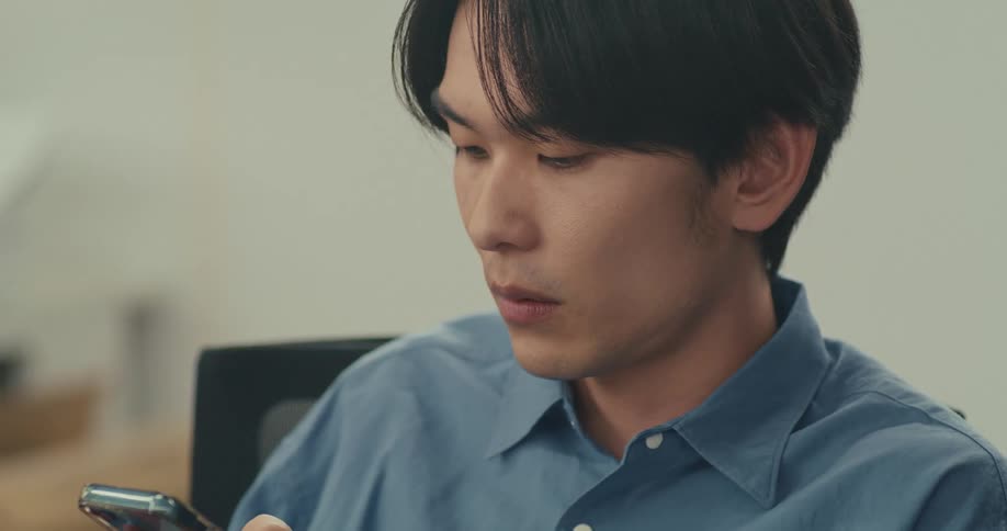 A close-up shot of a serious young Asian man scrolling intently on his smartphone indoors