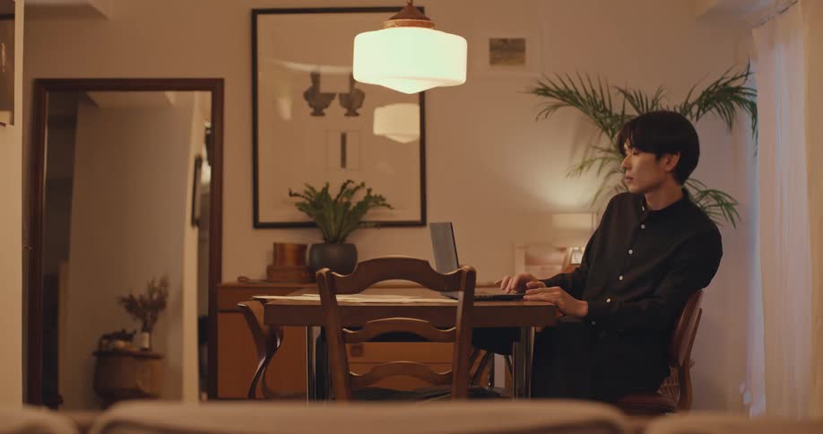 A young Asian man sits thoughtfully at his dining table at night, with his laptop open in a stylishly decorated, warmly lit room