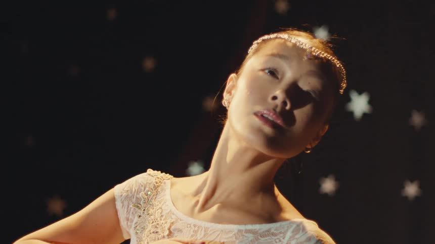 An elegant ballerina in a sparkling tiara and white leotard poses gracefully in slow motion on a stage with a starry background