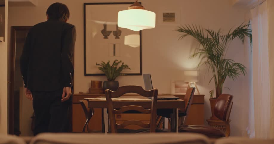 A young man, looking anxious, abruptly stands up from his laptop at the dining table and walks away at night