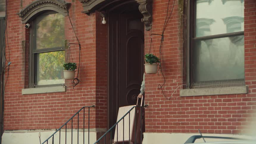 A young woman carries her suitcase up the front steps of a brick apartment building and goes inside, arriving home