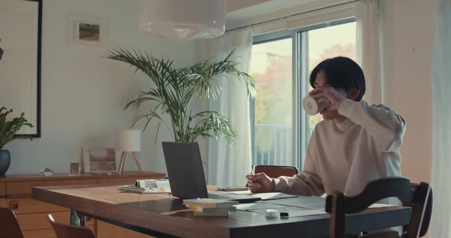A young Asian man works on his laptop and drinks from a mug while sitting at a dining table in his sunlit home