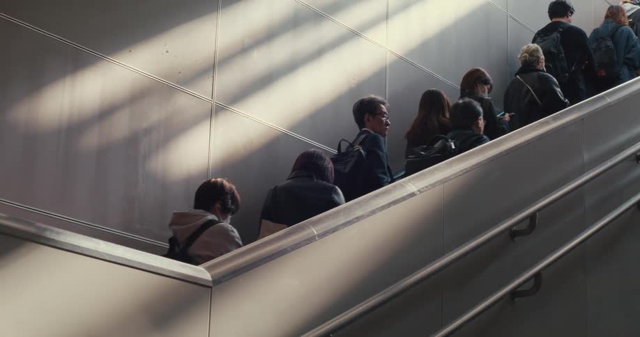 Anonymous commuters travel up a modern escalator, their journey highlighted by diagonal beams of sunlight