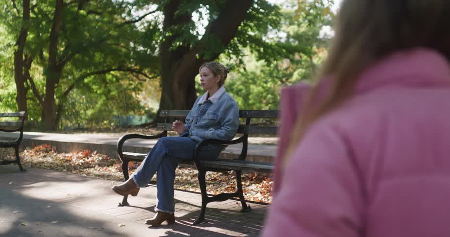 A young woman wearing a denim jacket sits alone on a park bench, looking up from her phone as she waits on a sunny autumn day