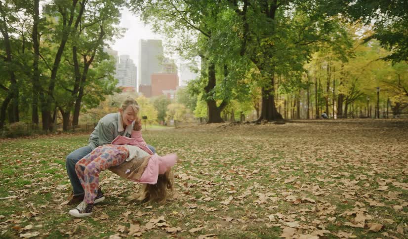 A mother and her young daughter laugh and play together in an autumn park, surrounded by fallen leaves with a city skyline in the background