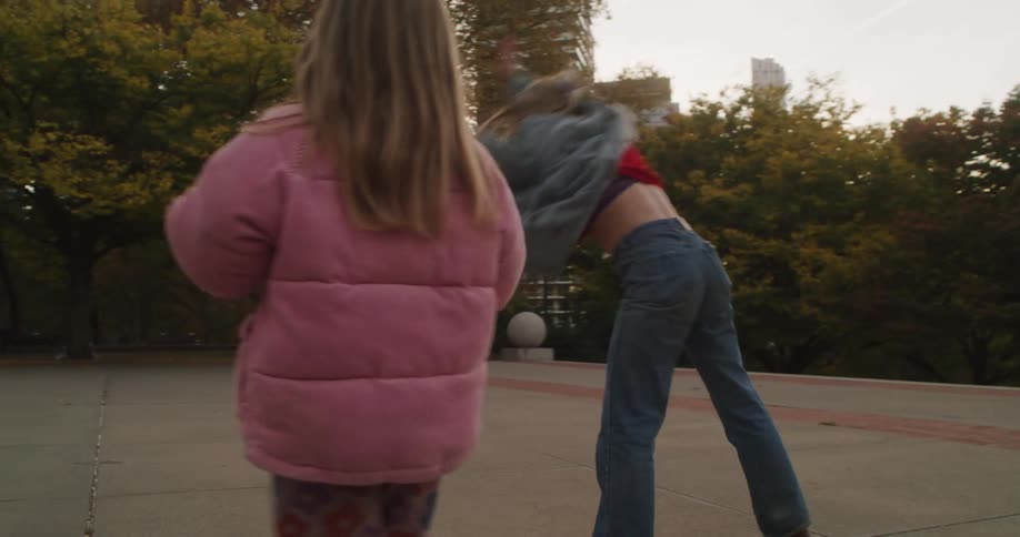 A young woman does a cartwheel in slow motion for a smiling little girl in a city park during autumn