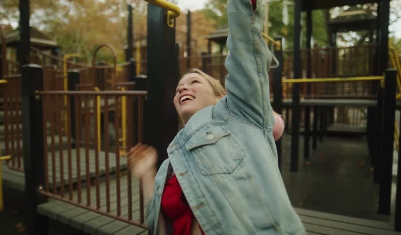 A playful woman smiles and hangs from the monkey bars in a park playground while a little girl watches her