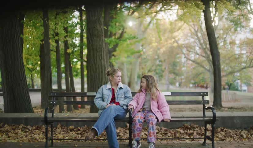 A little girl sits down next to her older sister on a park bench, holding her hand and having a conversation