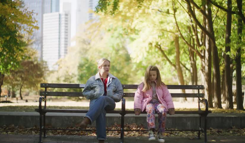 A young woman and a little girl sit and talk on a park bench on a sunny autumn day with city buildings in the background