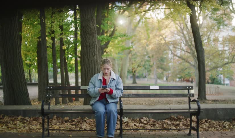 A sad and anxious young woman waits on a park bench in the autumn, checking her phone before looking up with disappointment