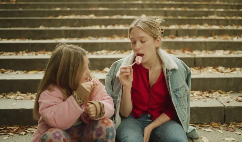 A young woman and a little girl sit together on park steps covered in autumn leaves while eating pink lollipops