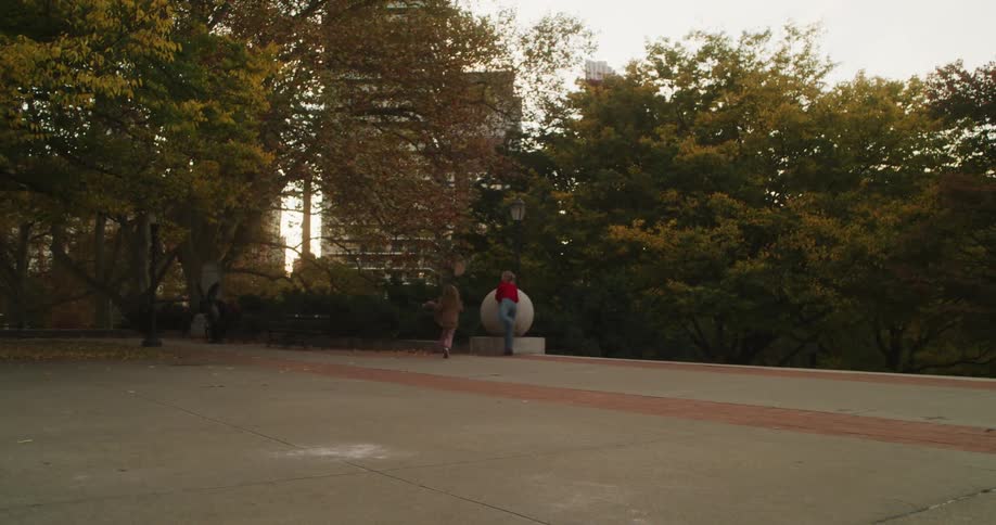 A mother and her young daughter playfully chase each other and dance in a city park on a beautiful autumn day