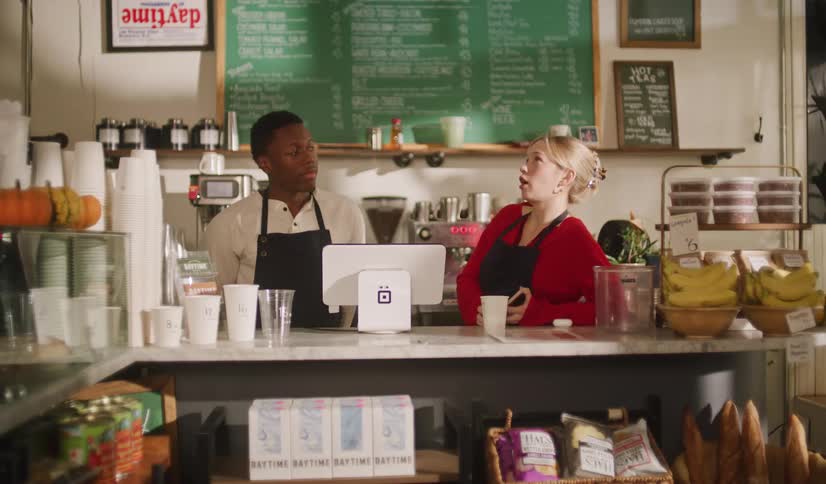 Two young baristas, a man and a woman, stand behind the counter of a cafe talking while waiting for the next customer