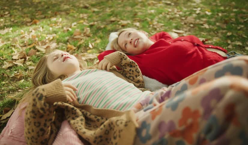 Two young girls lie on the grass among fallen autumn leaves, talking and enjoying the sunshine together