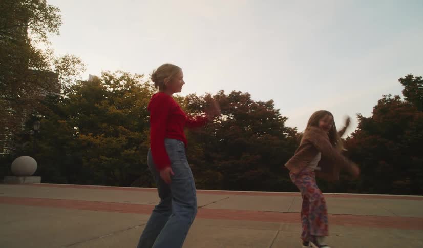 A happy mother and her young daughter dance energetically in slow motion, kicking and spinning with joy in a city park at dusk