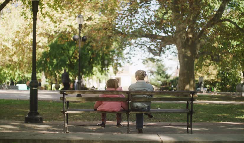 A woman and a young child sit on a park bench together, enjoying a peaceful autumn day as people walk by in the background