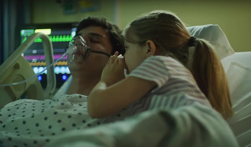 A worried young daughter whispers into her ill father's ear as he lies unconscious in a hospital bed