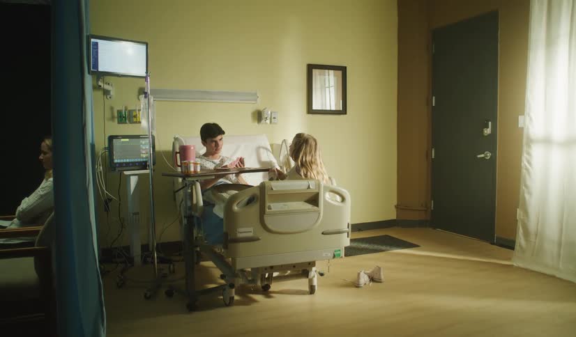 A young girl visits her friend in the hospital as his mother sits in a chair nearby
