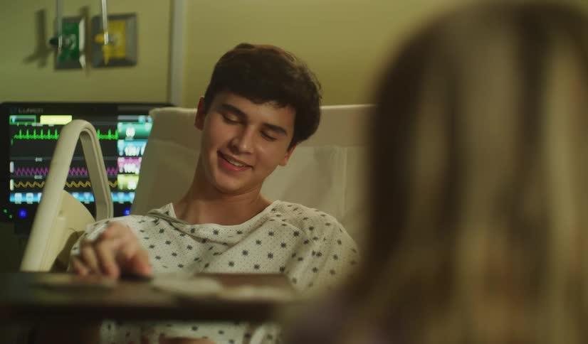 A teenage boy in a hospital gown plays cards with a friend from his hospital bed, his emotions shifting as they talk