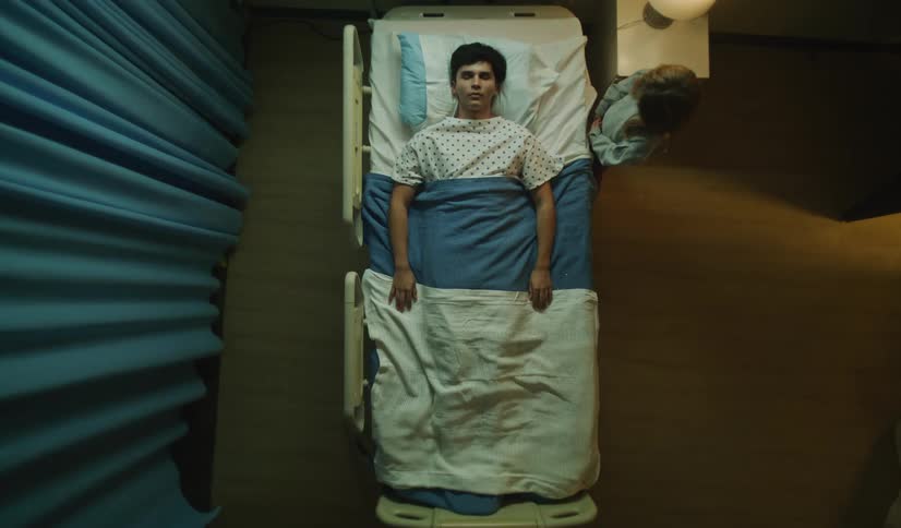 Overhead view of a solemn hospital room where a young man lies in bed as one visitor leaves and another remains seated