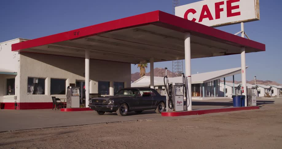 A classic Ford Mustang pulls up to a retro desert gas station where a woman gets out and then drives away