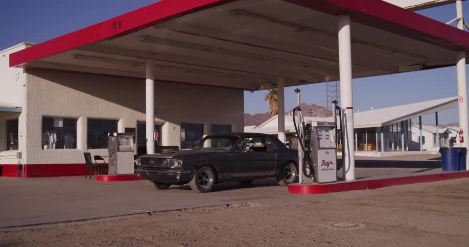 A woman driving a classic Ford Mustang pulls into a retro desert gas station to refuel on a sunny day