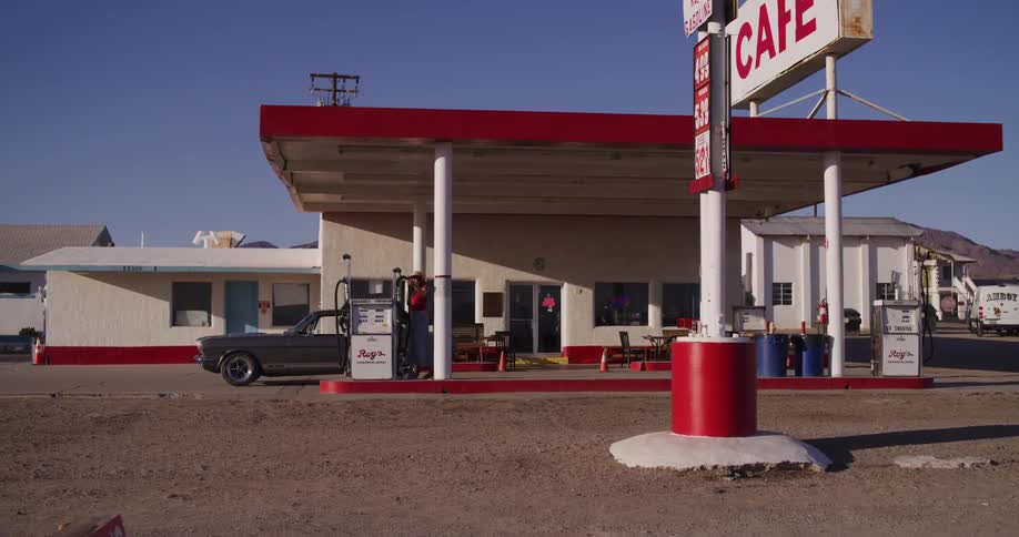 A woman in a cowboy hat pulls her classic Mustang into a retro roadside gas station in the desert to refuel