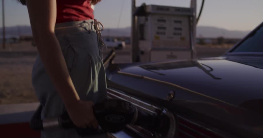 A young woman wearing a hat gets out of her classic car and refuels it at a remote desert gas station at sunset