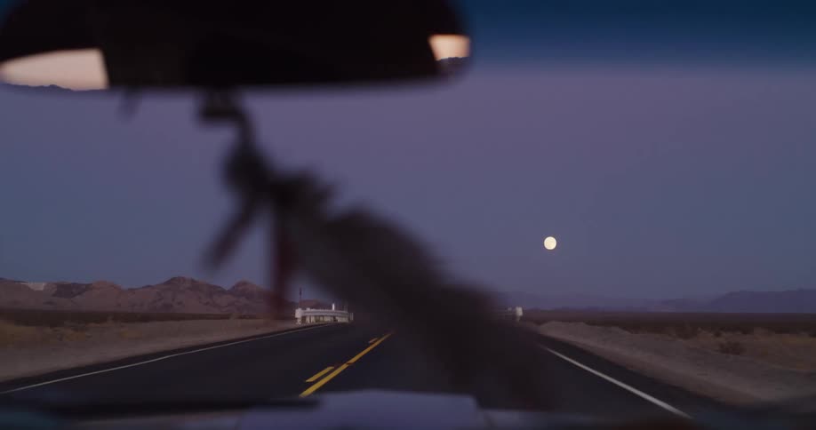 A POV shot from a car driving down a long desert highway at twilight, following the full moon on the horizon