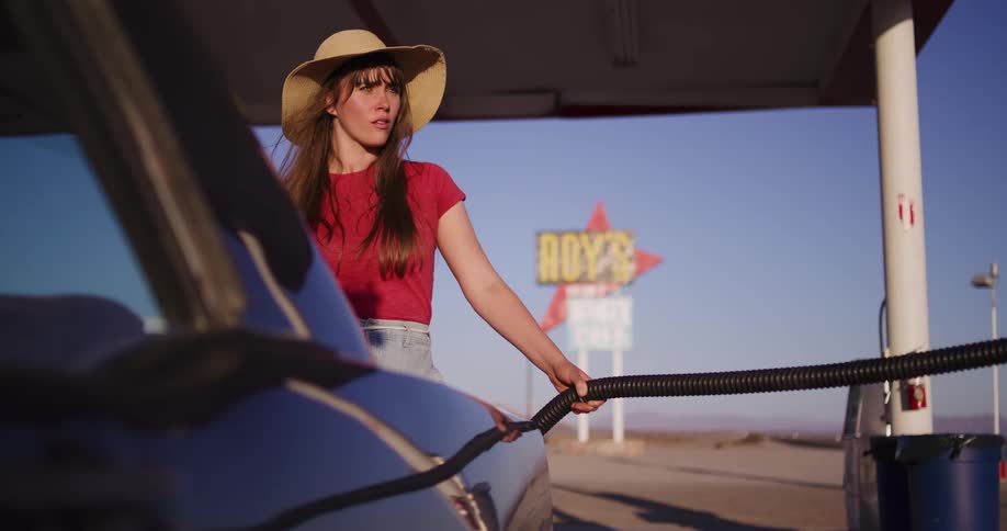 A woman in a hat refuels her car at a retro desert gas station as the sun sets