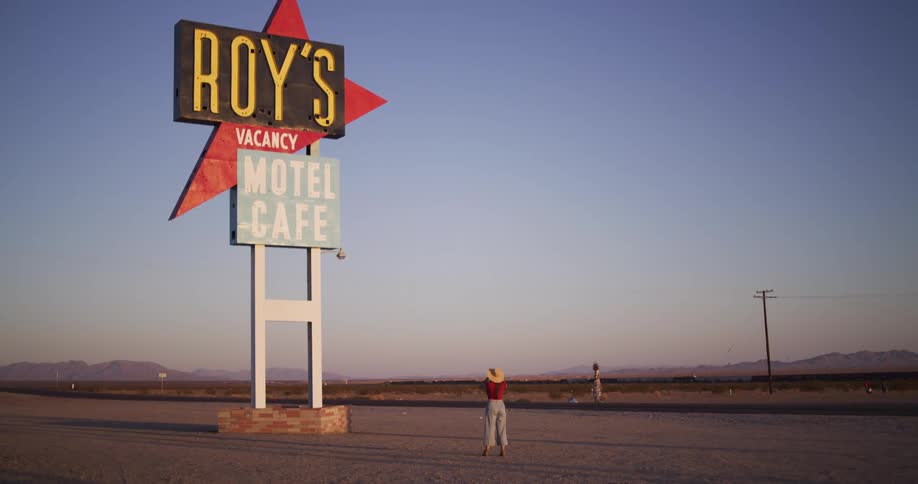 A lone woman wearing a hat walks up to the iconic vintage Roy's Motel Cafe sign along a desolate desert highway at sunset