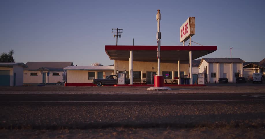 A vintage muscle car pulls up to refuel at a classic American roadside gas station and cafe before driving away
