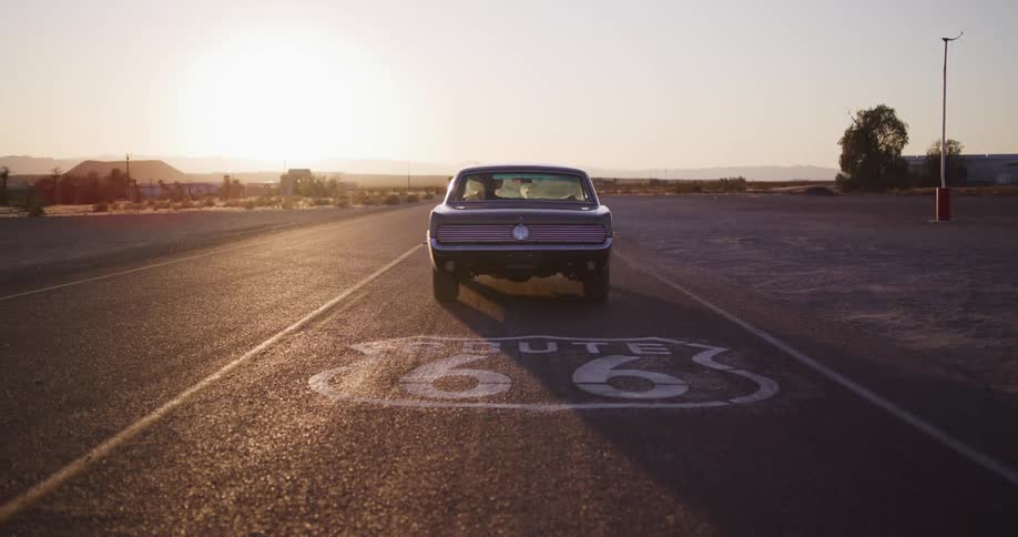 A woman in a classic car begins her American road trip, pulling away from a Route 66 sign and driving into the desert sunset