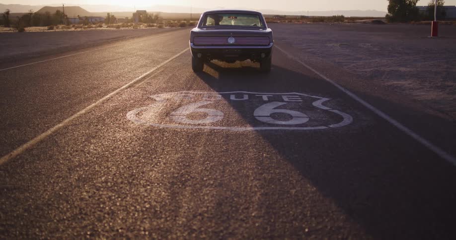 The camera pulls back from a classic Ford Mustang to reveal the iconic Route 66 road sign as the car drives off into the desert sunset