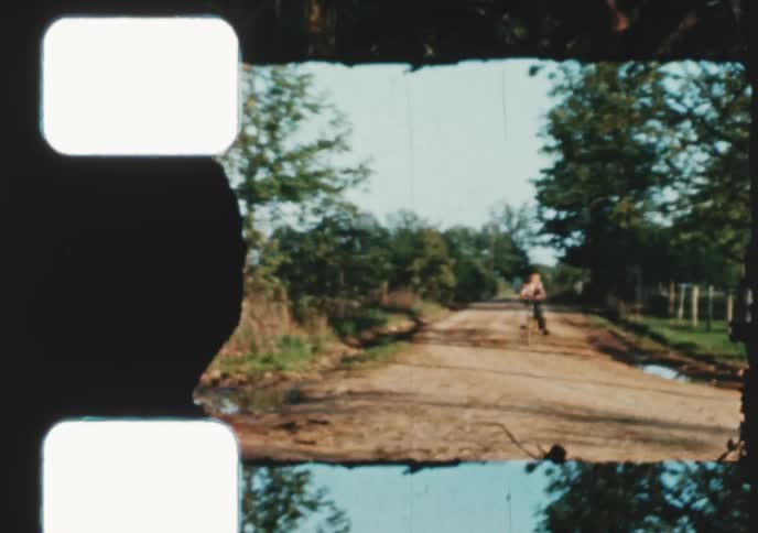 Vintage 8mm film shows a young girl and boy riding their bicycles down a rural dirt road in the summertime