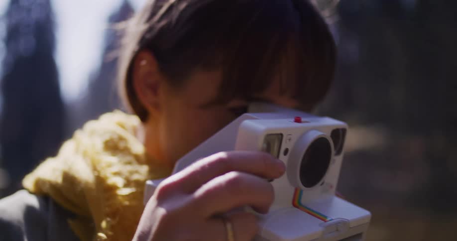 A smiling young woman in a yellow scarf captures a memory with a white instant camera in a sunlit forest