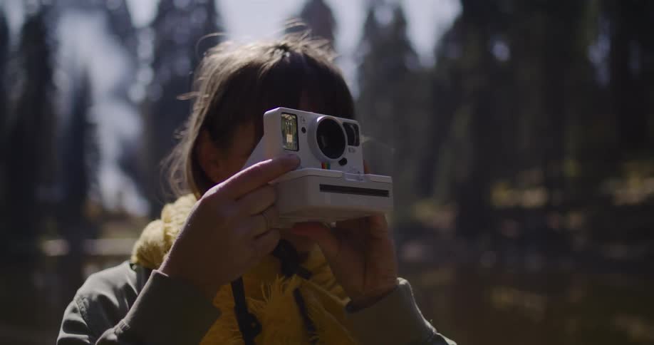 A young woman wearing a yellow scarf frames a shot and takes a picture with her instant camera in a beautiful forest setting