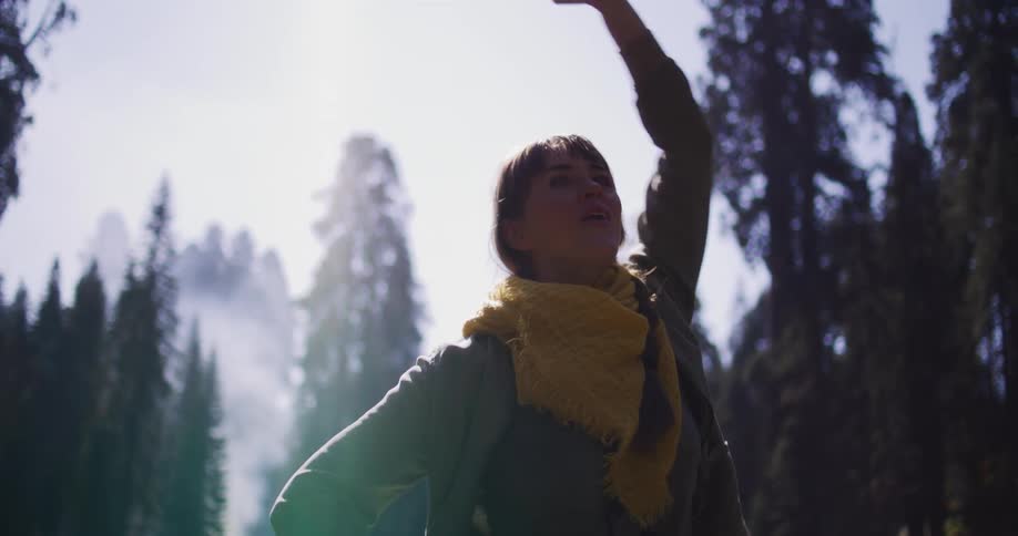 An awe-struck woman in a yellow scarf raises her hand to the sky in a sunlit, foggy forest in slow motion