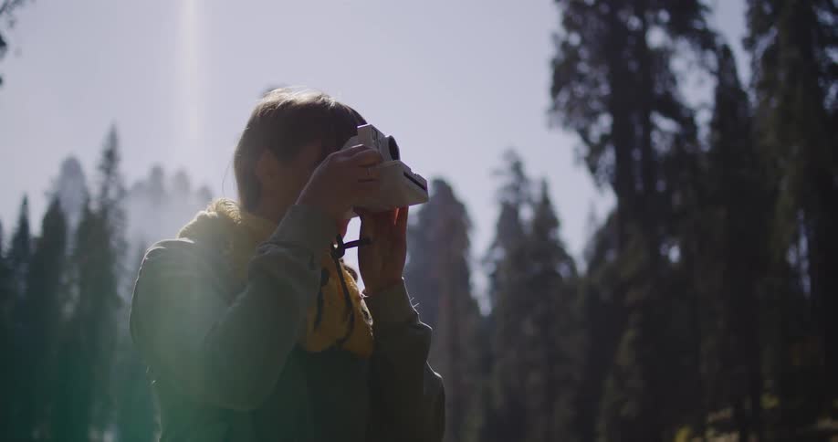A female photographer takes a picture of the majestic forest with a classic instant camera and then gazes at the scenery