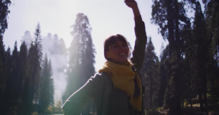 An adventurous woman triumphantly raises her arm in the air after running to a scenic spot by a lake in a sun-drenched, misty redwood forest