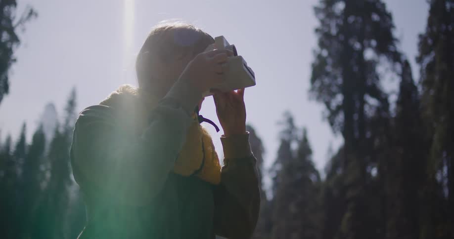 A young woman takes a photo with a vintage instant camera in a sun-drenched forest, capturing a moment on her adventure
