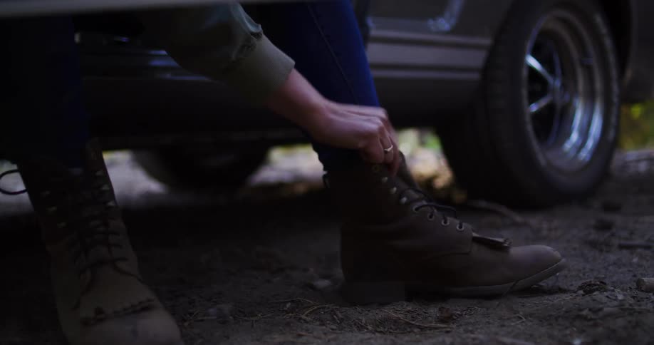 A woman sits in the open door of a classic car parked in a forest and ties her hiking boots