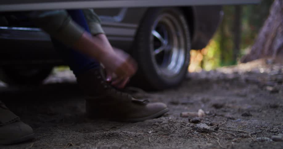 A person gets out of their classic car parked in a forest and sits down to tie their brown hiking boots