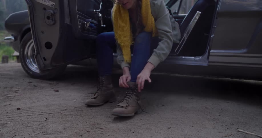 A woman sits in the doorway of a classic car parked in a forest and carefully ties her hiking boots