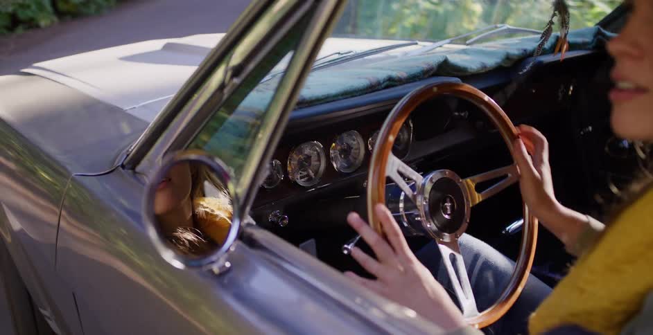 A young woman enjoys a drive in her classic convertible, her reflection visible in the side mirror as she holds the wooden steering wheel