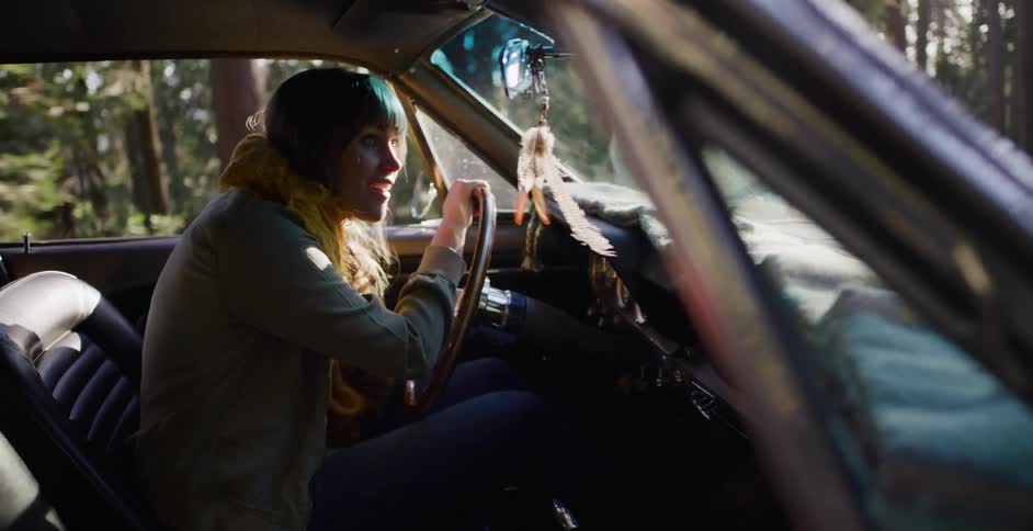 A young woman sits in the driver's seat of a classic car, talking happily while parked in a sun-dappled forest